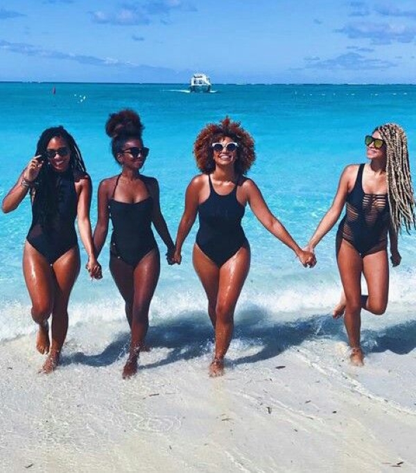Quatro mulheres de pele negra usando maiô preto de mãos dadas saindo do mar
