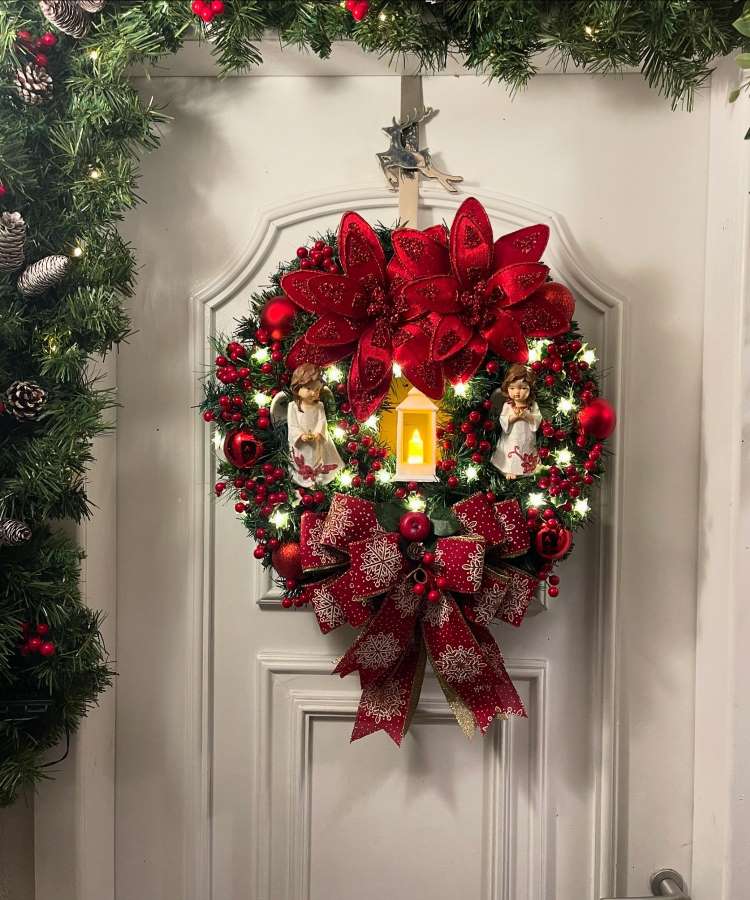 Enfeite de Natal de porta exuberante com grandes flores vermelhas e laço decorativo. Adornado com pequenas luzes, bolas e bagas vermelhas. Dois anjos brancos flanqueiam uma vela central, criando uma cena angelical sobre uma porta branca, cercada por folhagens verdes.
