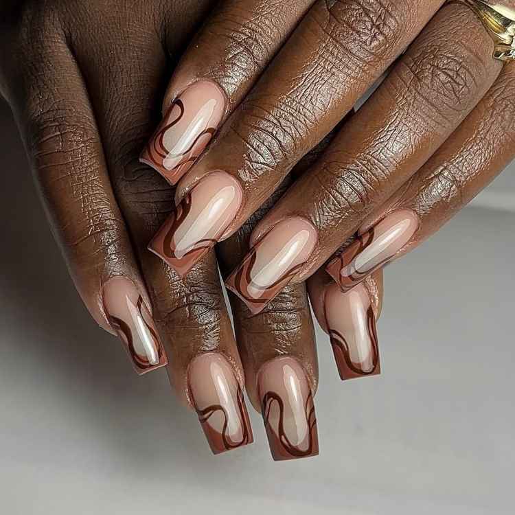 Unhas longas, quadradas, decoradas com esmalte nude e bordas em ondas marrons e brancas. O visual combina elegância e modernidade, com acabamento brilhoso. As mãos estão sobrepostas contra um fundo claro, destacando o contraste entre o design e a pele.