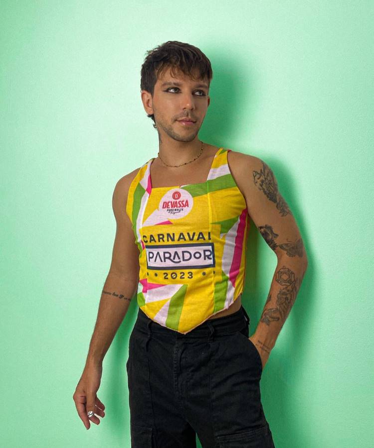 A imagem mostra um homem posando com um abadá customizado em tons vibrantes de amarelo, verde, rosa e branco. O abadá traz as inscrições "Devassa", "Carnaval Parador 2023". Ele combina com calça preta, acessórios discretos e maquiagem marcante.