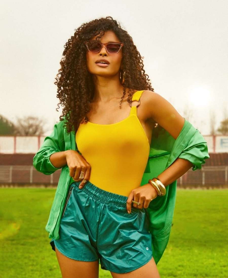 Imagem com fundo de campo de futebol. No centro, mulher usando roupas Brazilcore: camiseta amarela + short verde + camisa verde.