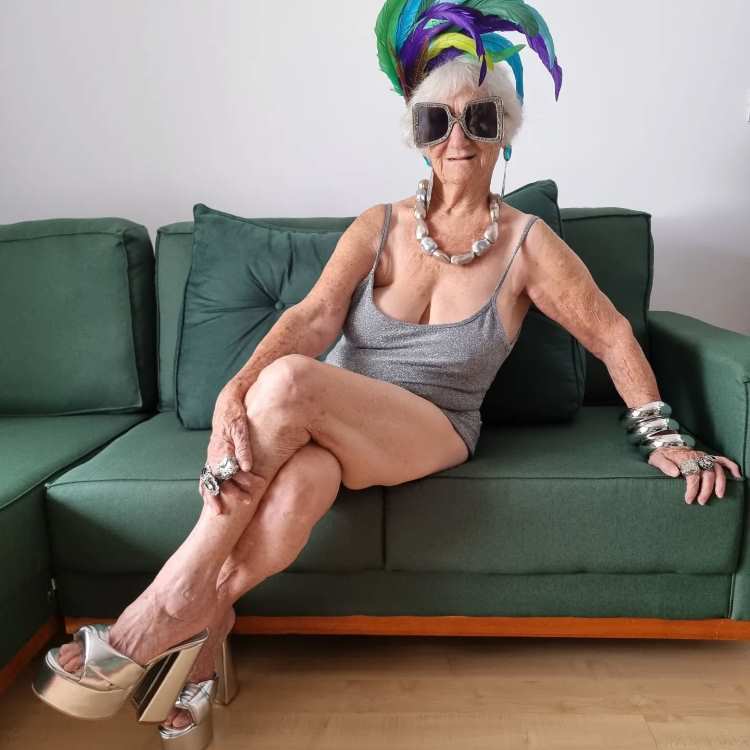 Mulher idosa estilosa sentada em sofá verde, veste maiô prateado, salto alto dourado e acessórios chamativos, incluindo colar, pulseiras e anel. Usa óculos escuros grandes e tiara com penas coloridas, transmitindo confiança e originalidade em ambiente descontraído.