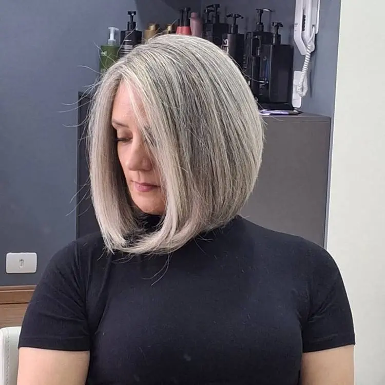 Uma mulher de cabelos grisalhos curtos e lisos, com corte bob assimétrico, inclina a cabeça para baixo. Ela veste uma blusa preta e o fundo mostra prateleiras com produtos de beleza.