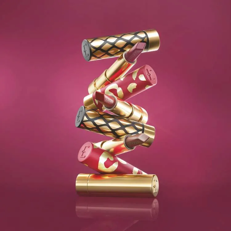 Vários batons Eudora empilhados artisticamente, com embalagens douradas e decorativas, em um fundo rosa. As cores variam entre tons de vermelho e nude. Modelo do desfile da Neriage patrocinado pela Eudora.