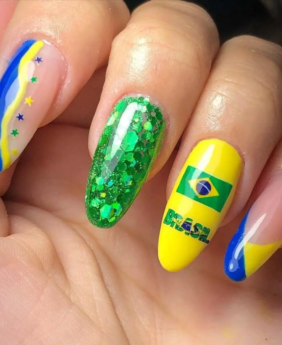 Imagem de mão decoradas com esmalte. Cores usadas, verde, amarelo e azul. Indicador: transparente com azul, amarelo e estrelas. Dedo Médio, glitter verde. Anelar: amarelo com bandeira do Brasil e escrito "Brasil". Mínimo: transparente com francesinha azul e amarelo.