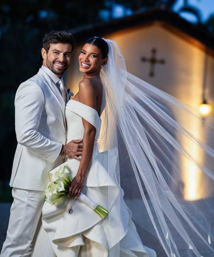 Casal Jakelyne Oliveira e Mariano posa sorridente durante casamento ao ar livre. Noiva veste vestido branco volumoso, com véu longo esvoaçando, e segura buquê de lírios. Noivo usa terno claro, destacando elegância. Ao fundo, capela iluminada com cruz acima da entrada.
