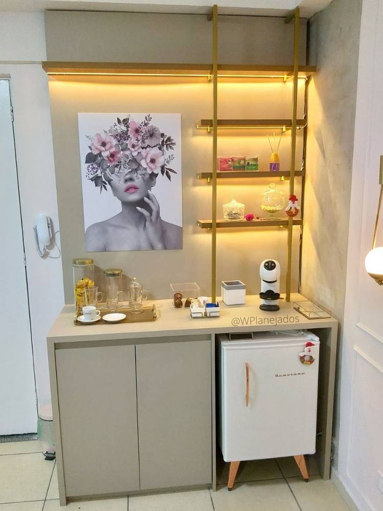 Cantinho do café iluminado, com frigobar, quadro com flores, prateleiras, xícaras e muito mais