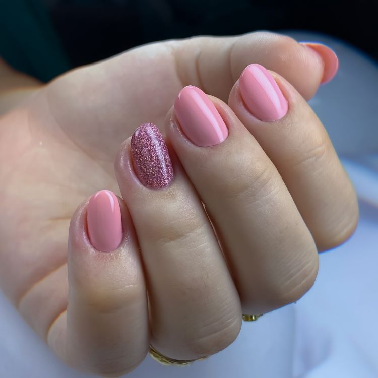 Mão exibindo estilos de unhas curtas, pintadas em esmalte rosa claro, com destaque para uma unha decorada com glitter rosa brilhante. Fundo desfocado em tons suaves de azul e branco, enfatizando o visual delicado, feminino e moderno das unhas.