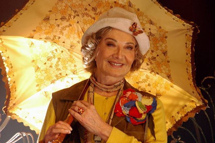 A atriz mais jovem atuando para novela, usando roupa colorida, chapéu e guarda-chuva florido