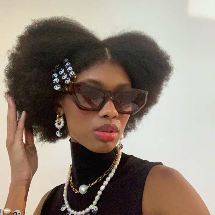 Mulher com cabelo afro volumoso repartido ao meio, decorado com presilhas de pérola, óculos escuros grandes e colares de pérolas, usando blusa preta sem mangas.