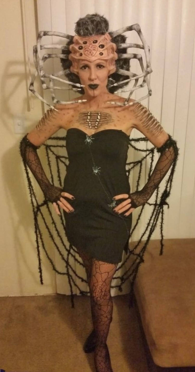 Mulher de pele clara fantasiada de aranha, com adereço que imita aranha na cabeça, vestido preto e capa de teia 