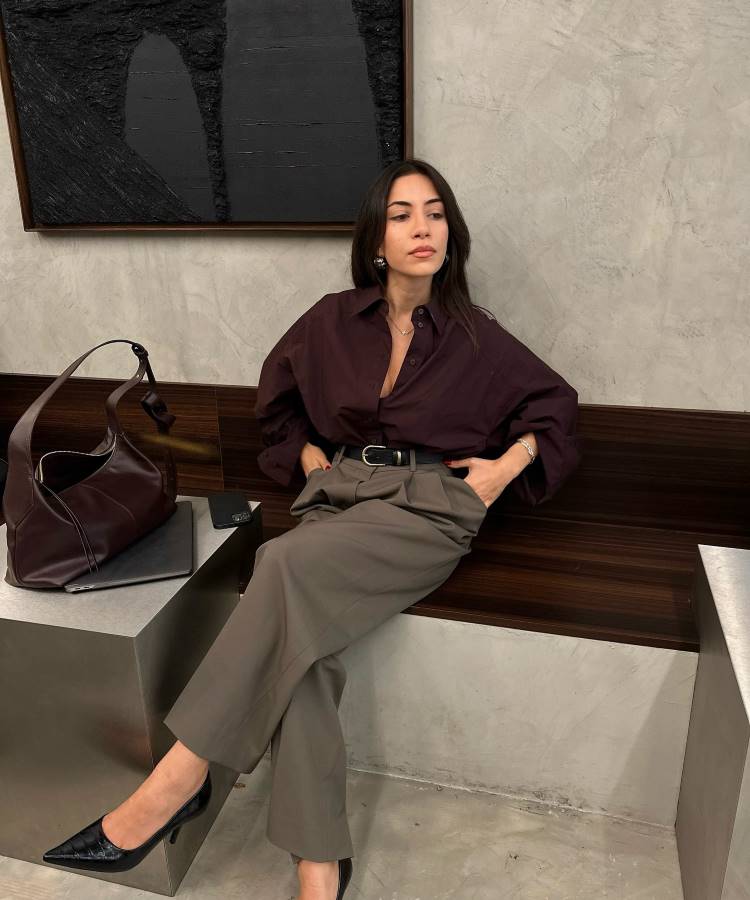 Mulher elegante sentada em banco de madeira, usando camisa vinho oversized, calça bege com cinto preto e sapatos de salto pretos. Ao lado, bolsa estruturada marrom sobre mesa metálica. Inspiração moderna e sofisticada para looks de trabalho profissionais.





