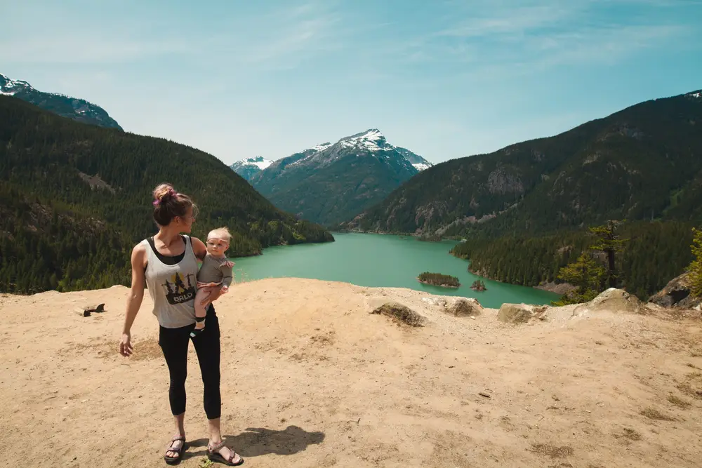 Mãe de pele clara com um bebê no colo no meio da natureza, com montanhas e rio.