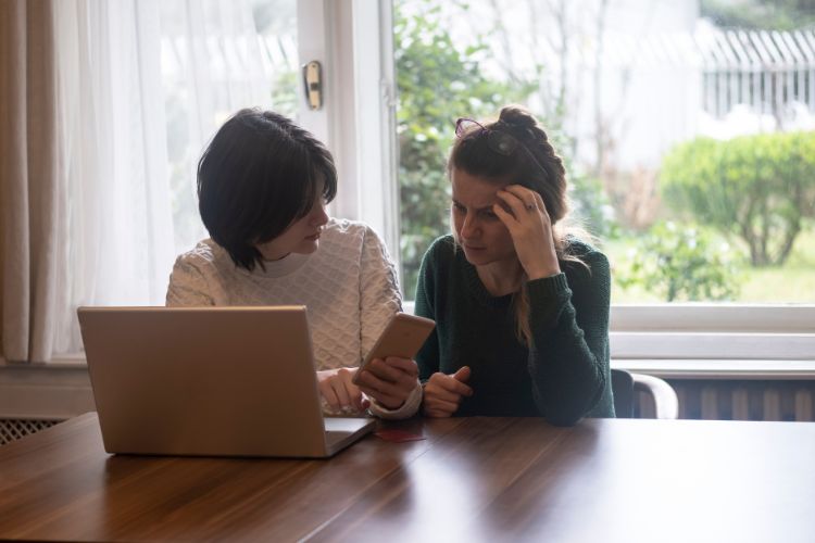 Duas mulheres pensativas segurando celular e notebook sentadas em uma mesa com janela no fundo sofrendo golpe por inteligência artificial