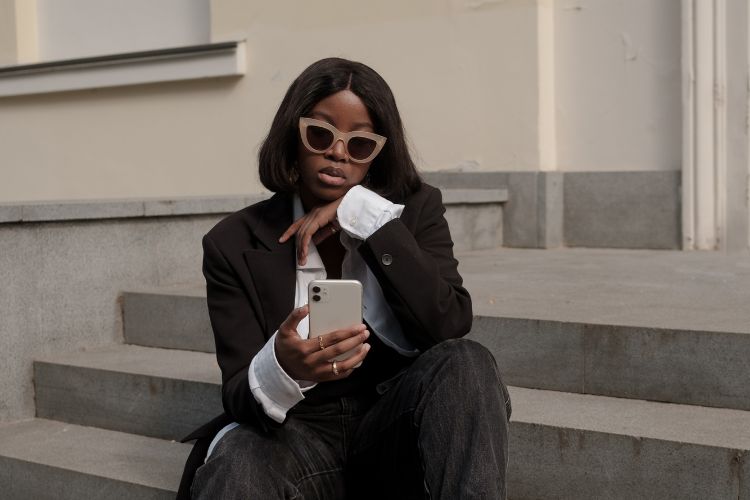 Mulher sentada em degraus, usando blazer preto, camisa branca e óculos de sol elegantes. Ela olha para seu celular com pose reflexiva, possivelmente pesquisando temas como “Quanto custa um publi?” ou analisando estratégias de conteúdo para redes sociais.
