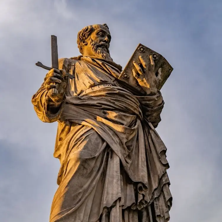 Uma estátua de pedra desgastada de Paulo Apóstolo se destaca contra um céu dramático. Ele segura uma espada e um livro, símbolos frequentemente associados a ele, retratado com uma túnica fluida e barba.