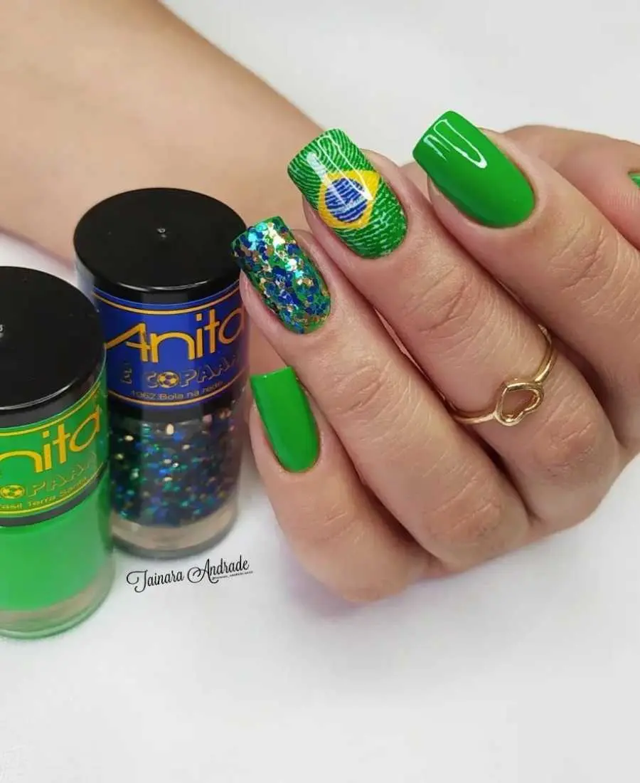 Imagem de mão decoradas com esmalte.  Cores usadas, verde, amarelo, azul e branco. + glitter azul e dourado. Arte, desenho de bandeira no dedo médio, todas unhas com fundo verde e unha do dedo anelar com os glitteres espalhados.