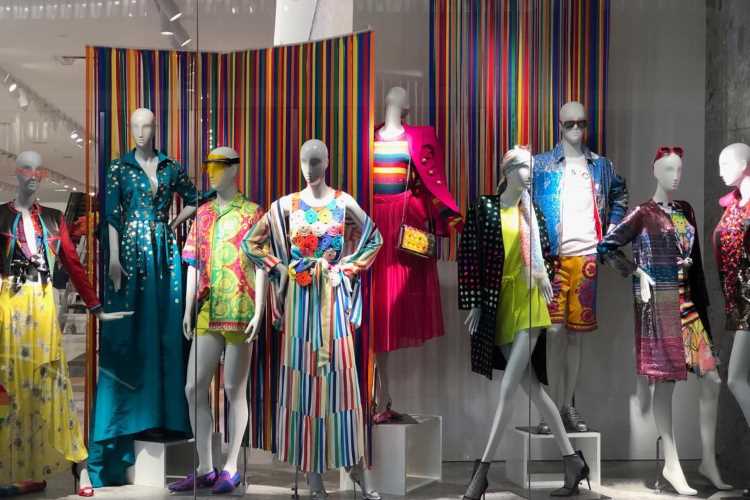Vitrine com manequins exibindo roupas vibrantes e estampadas, destacando listras, flores e padrões geométricos. Painéis coloridos ao fundo complementam o visual. A iluminação realça os detalhes contemporâneos e ousados, criando uma atmosfera moderna e artística de moda.
