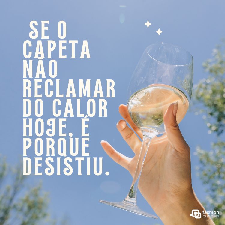 Imagem com o céu azul ao fundo, uma mão segurando taça de vinho branco em destaque e vegetação ao redor. Texto humorístico diz: “Se o capeta não reclamar do calor hoje, é porque desistiu.” Enquadrado em frases de calor.