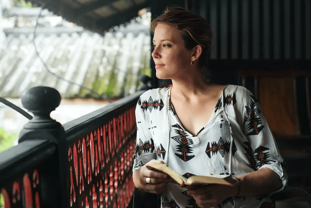 Mulher de pele clara usando blusa branca na sacada com livro na mão