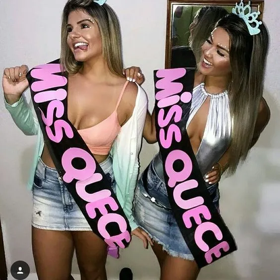 Duas mulheres de pele clara com faixa preta escrito "miss quece" por cima da roupa 