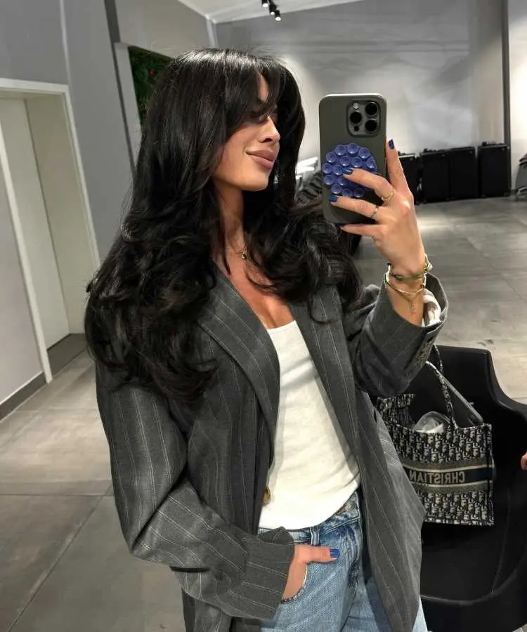 Mulher com cabelos longos, pretos e volumosos, estilizados em camadas, usando blazer cinza risca de giz, blusa branca e jeans. Ela tira uma selfie com expressão confiante em um ambiente moderno, com detalhes neutros e iluminação suave.