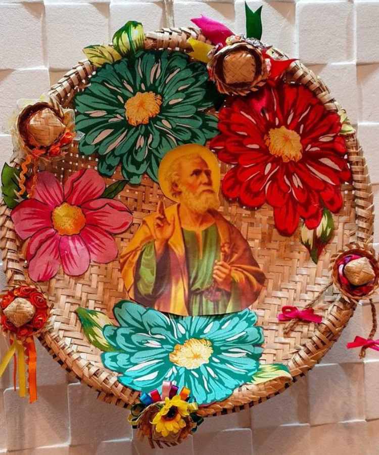 A imagem representa uma peneira de palha decorada com imagem de São João e minis chapéus de palha