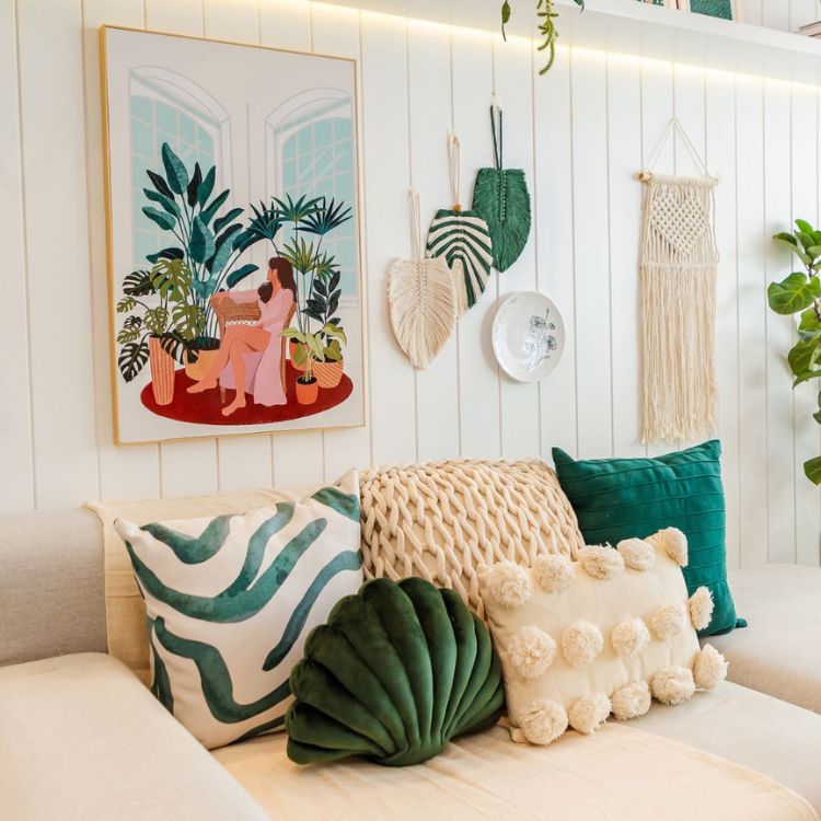 Sala com parede branca, quadro de planta, macramês pendurados, sofá bege e almofadas de diferentes cores, formatos e texturas, uma ideia de decoração barata