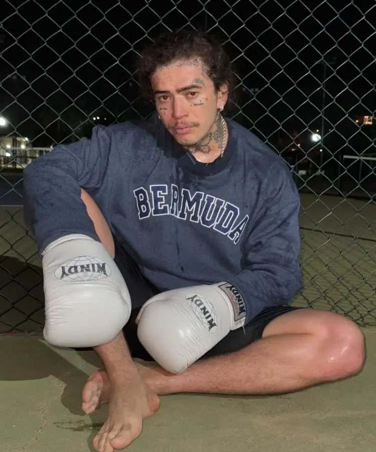 A imagem mostra um homem sentado no chão, descalço, usando luvas de boxe brancas e uma blusa azul com "BERMUDA" estampado. Ele tem tatuagens no rosto e pescoço, com uma expressão séria, cercado por uma grade à noite.