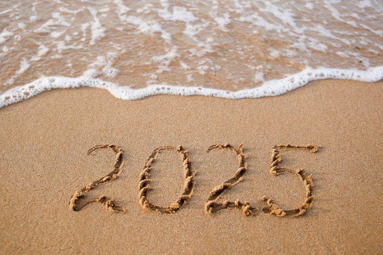 A imagem mostra a palavra "2025" escrita na areia de uma praia. Ondas suaves se aproximam, quase tocando a inscrição. O cenário transmite uma sensação de transição, renovação e conexão com a natureza, destacando o ano novo.