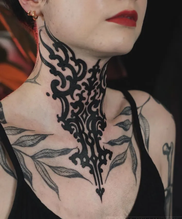 A imagem mostra uma pessoa de frente com cabelo curto e batom vermelho. Ela tem uma tatuagem no pescoço que se estende até o peito, com um design intrincado e simétrico em preto. A pessoa usa brincos pequenos e uma blusa preta.

