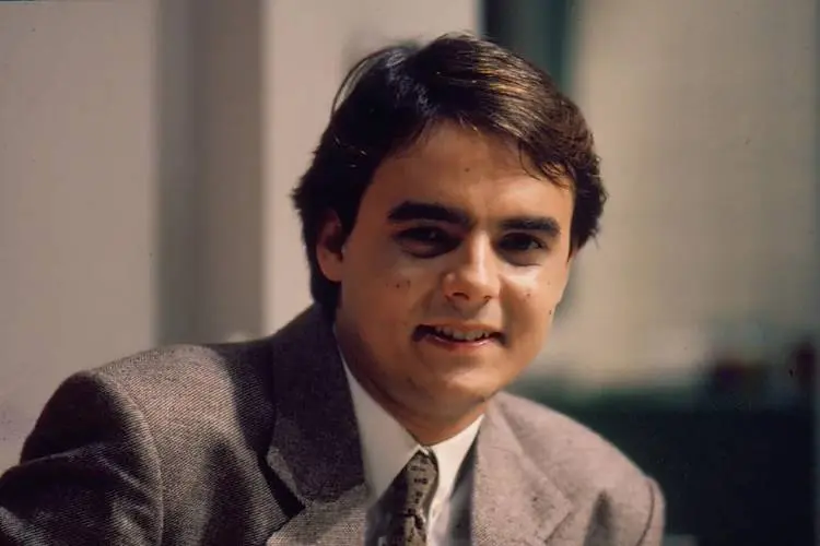 Homem jovem, usando terno cinza e gravata estampada, com cabelos castanhos e sorriso discreto. Ambiente interno com fundo desfocado. Figura que integrou o elenco de "Vale Tudo" hoje é relembrada por seu papel marcante na novela.