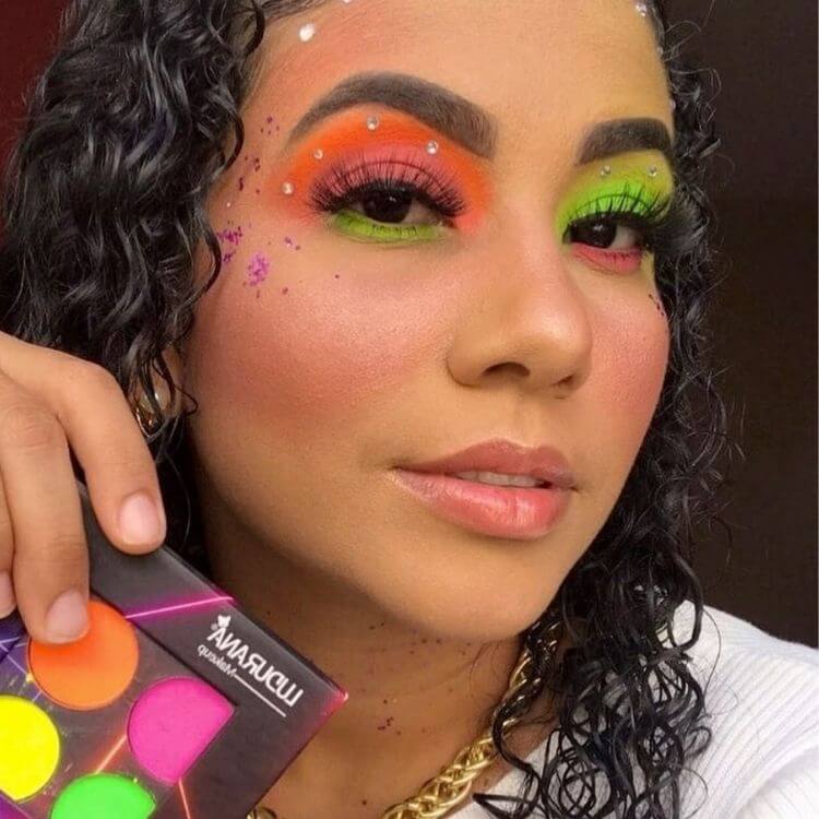 Mulher de pele morena usando sombras neon em laranja, rosa e verde, além de glitter roxo e strass
