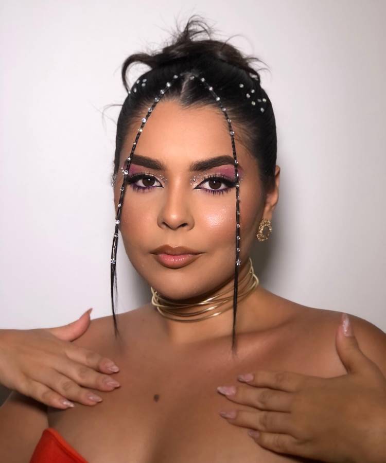 A mulher exibe um penteado elegante com coques altos e tranças finas decoradas com pedrinhas brilhantes. Sua maquiagem destaca tons de rosa e brilho, complementada por brincos dourados, um colar moderno e um vestido vermelho vibrante.