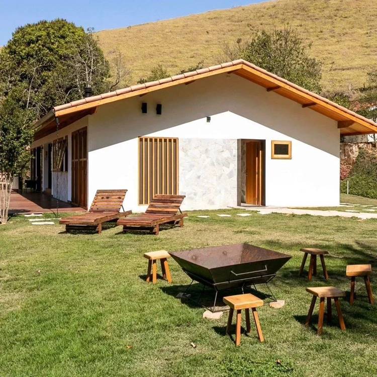 Imagem de fachada de casa simples branca, com jardim de grama, espreguiçadeiras de madeira, bancos de madeira e lareira de ferro