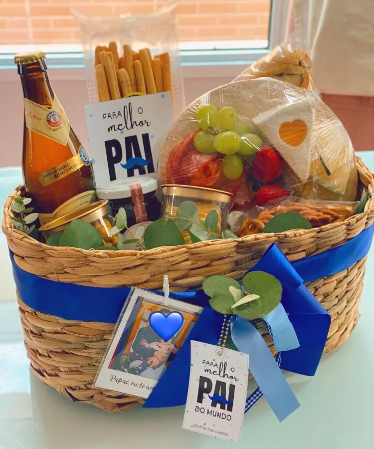 A imagem mostra uma cesta de Dia dos Pais com itens gourmet, incluindo uma garrafa de bebida, palitos crocantes, frutas frescas e embalagens de alimentos. A cesta é decorada com laços e cartões temáticos que expressam amor e apreço, como “Para o melhor PAI do mundo”.

