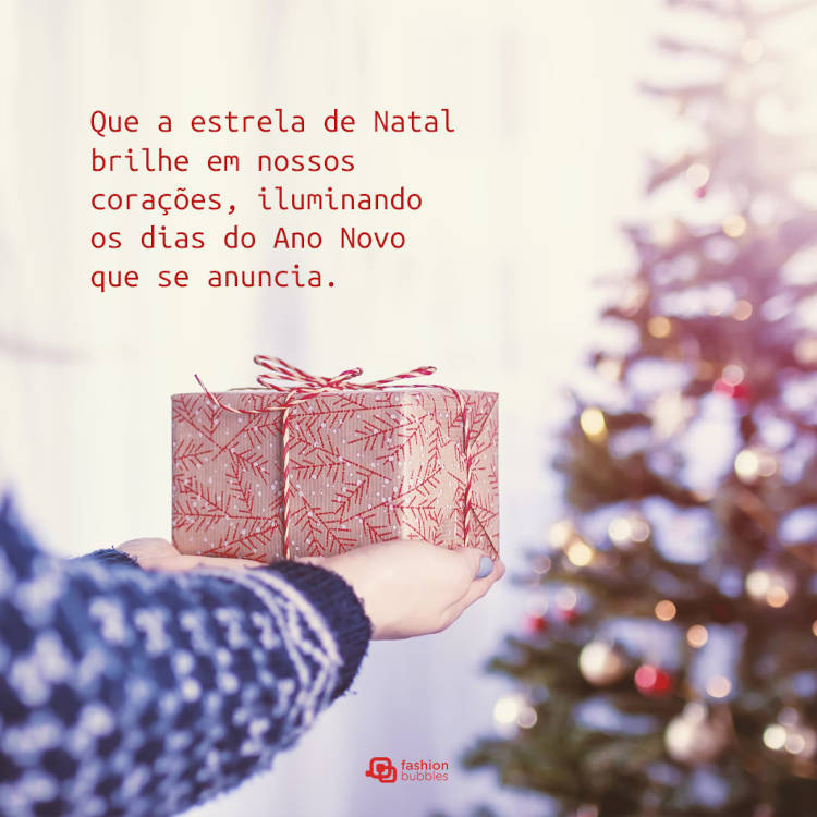 Pessoa de suéter azul segurando embrulho em frente árvore de Natal e frase: Que a estrela de Natal brilhe em nossos corações, iluminando os dias do Ano Novo que se anuncia.