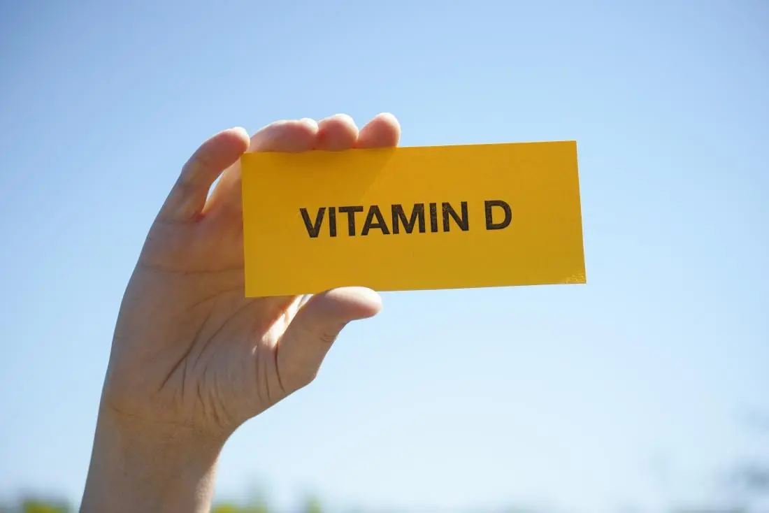 Mão de uma pessoa de pele clara segurando um papel amarelo escrito "VITAMIN D" em preto.