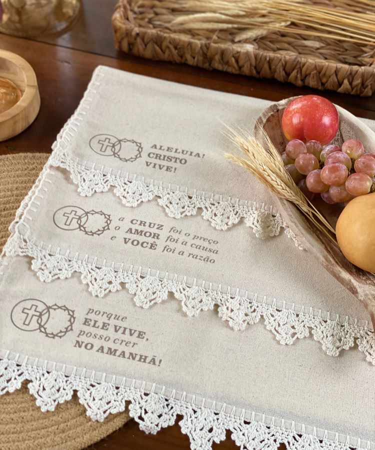 Mesa decorada com toalhas bordadas em crochê com mensagens cristãs: “Aleluia! Cristo Vive!” e outras frases. Ao lado, cesta de rattan com trigo, uvas, maçã e pão, compondo um ambiente rústico e delicado para a celebração religiosa.