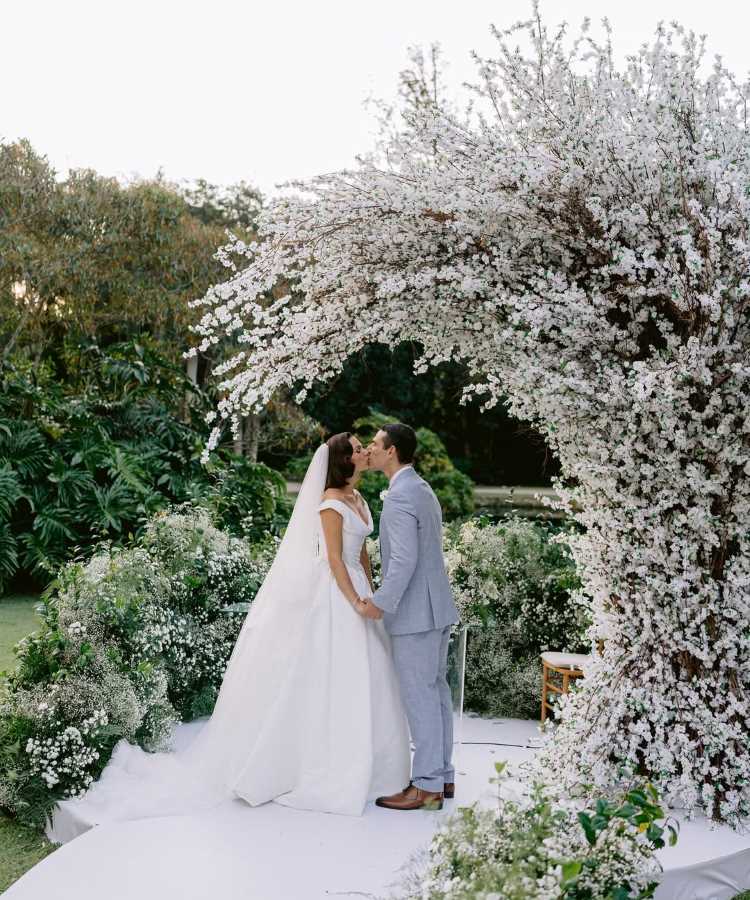 Casal Isis Valverde e Marcus Buaiz troca beijo romântico sob arco de flores brancas em cerimônia ao ar livre. Vestido de noiva clássico, traje azul claro do noivo e decoração exuberante destacam o ambiente idílico, com folhagens e flores criando um cenário encantador e elegante.
