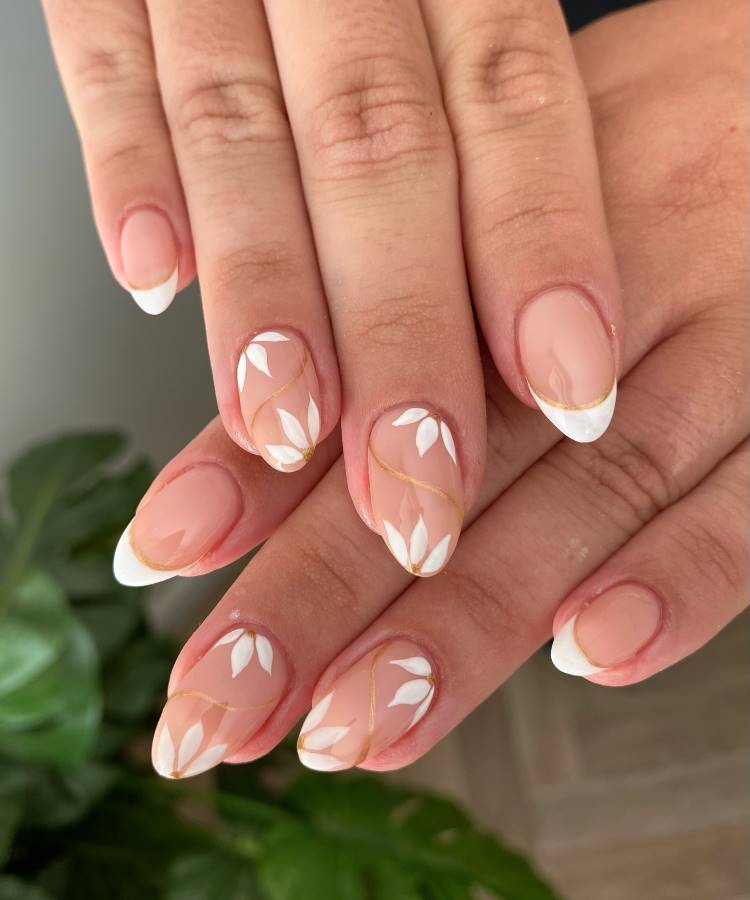 Mãos femininas com unhas ovais decoradas com esmalte nude, design francês branco e detalhes delicados de folhas brancas e linhas douradas. O fundo desfocado com plantas verdes cria um visual fresco, elegante e natural, destacando o estilo refinado.