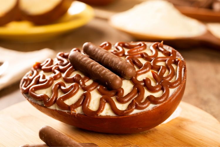 Metade de um ovo de chocolate recheado com creme branco, decorado com ganache de chocolate e dois chocolates em formato de palito no topo. O ovo está sobre uma tábua de madeira, com um fundo desfocado de cozinha.