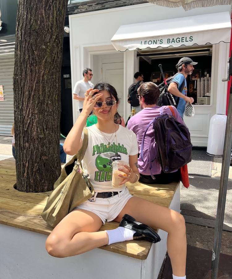 Uma mulher de cabelo escuro, usando uma camiseta branca com estampa e **shorts hot pantes verão 2026** brancos. Ela está sentada em um banco de madeira, com uma bebida na mão e óculos escuros, em frente a uma loja de bagels.