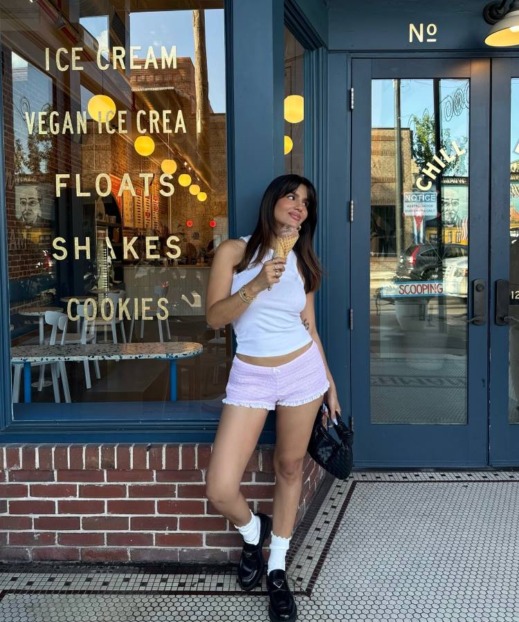 Mulher sorrindo, usando regata branca e shorts lilás. Ela segura um sorvete e posa em frente a uma loja. Usa meias brancas, um **sapato preto** tipo mocassim, e segura uma bolsa preta.