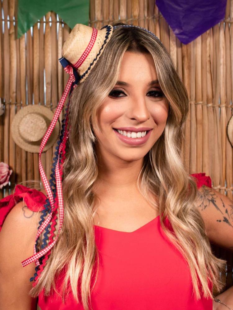 Mulher loira de pele clara usando tiara de festa junina, colorida e com chapéu, além de vestido vermelho