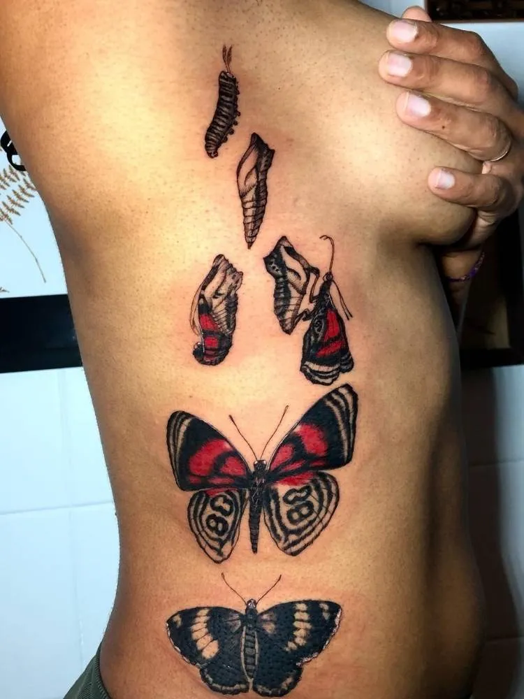 Tatuagem grande na costela da metamorfose de uma borboleta. Próximo ao seio, há uma lagarta no casulo que vai se tornando borboleta conforme a tattoo se aproxima da cintura. A tatuagem é preta e vermelha, em estilo mais realista e feita em uma mulher de pele morena.  
