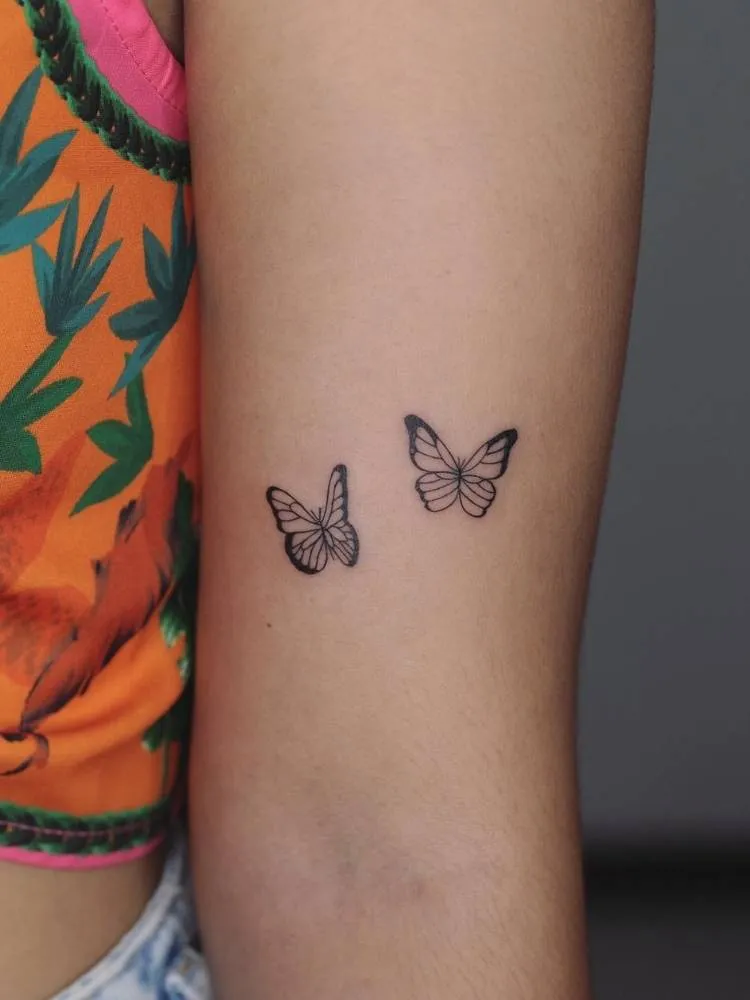 Tatuagem de duas borboletas pequenas, delicadas com traço fino no interior do braço de uma mulher de pele clara e vestindo uma blusa laranja e verde.