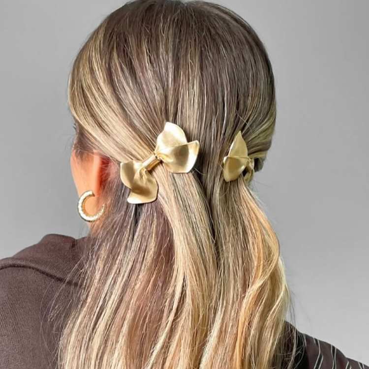 Cabelo loiro liso em penteado semipreso com presilhas douradas em formato de laços, combinando com brincos dourados.