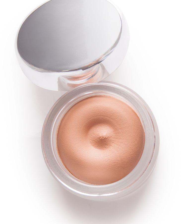 A imagem mostra um pote aberto de base em mousse, com uma textura suave e cremosa em tom bege. A tampa prateada está ao lado, refletindo a luz. Este é um dos tipos de base que oferece cobertura leve e acabamento natural, ideal para peles que buscam um efeito aveludado.


