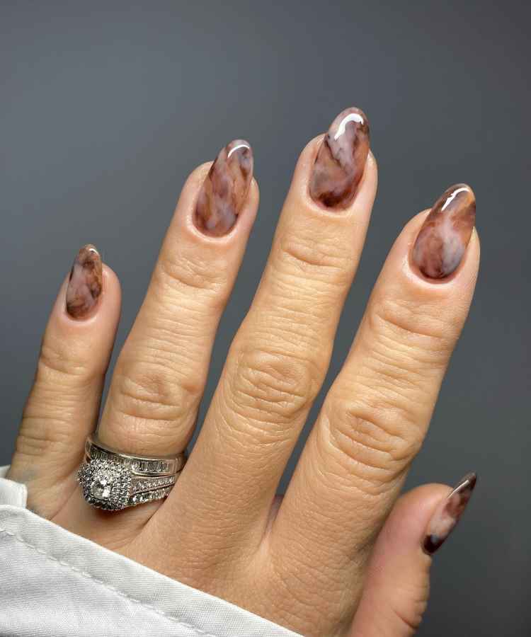 Unhas longas com esmalte marmorizado em tons de marrom e nude, criando um efeito sofisticado. A mão exibe anéis prateados com pedras brilhantes, destacando o visual elegante. O fundo cinza neutro realça o contraste e a beleza das unhas.